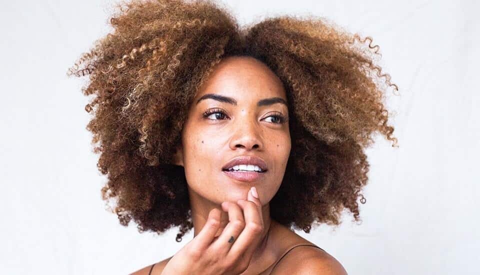A woman with afro hair is posing with her hand on her chin, showcasing her flawless skincare.